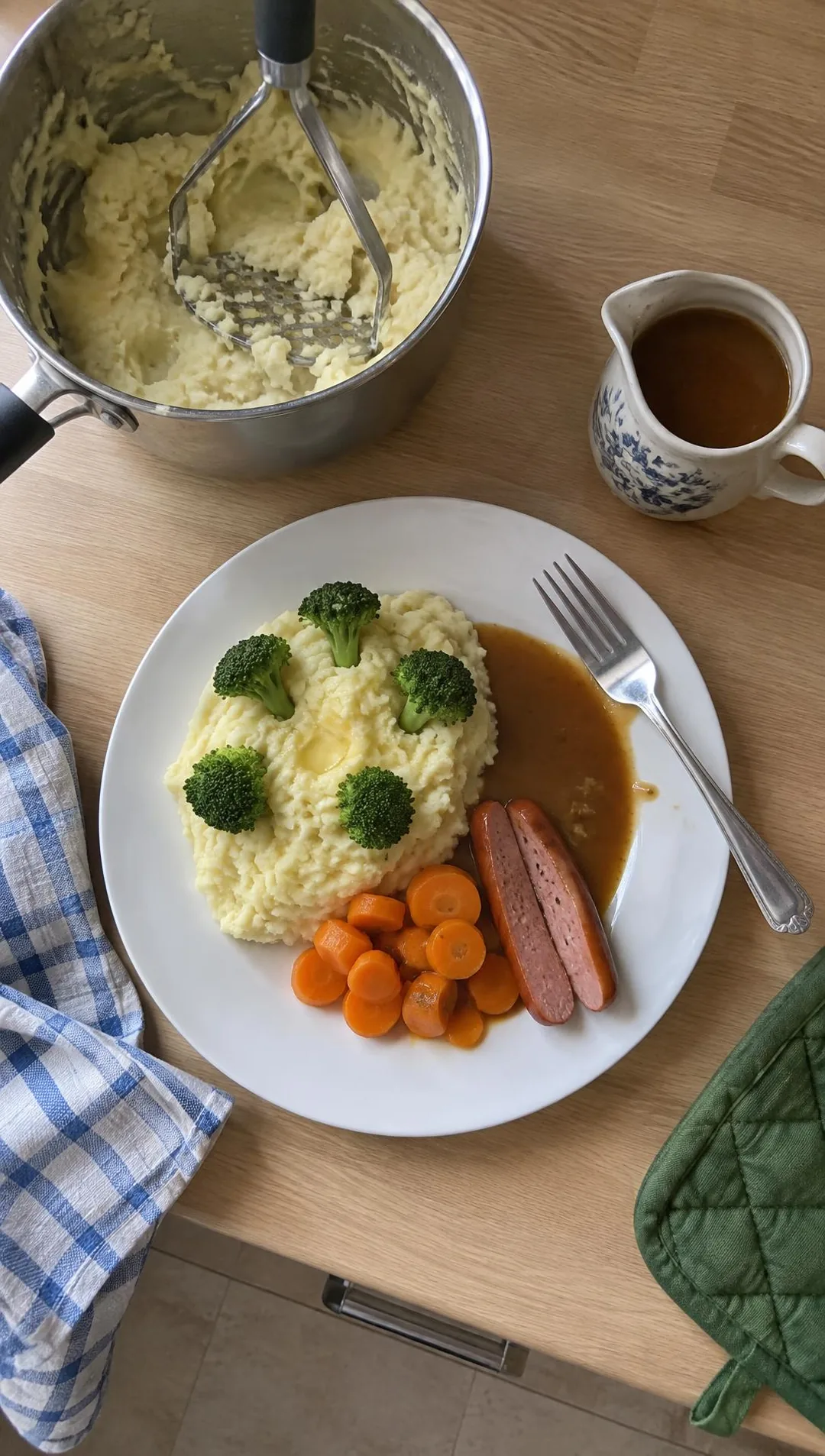 Teller mit Kartoffelbrei, darin Brokkoliröschen als Bäumchen, daneben Möhren, ein halbiertes Würstchen und Soße