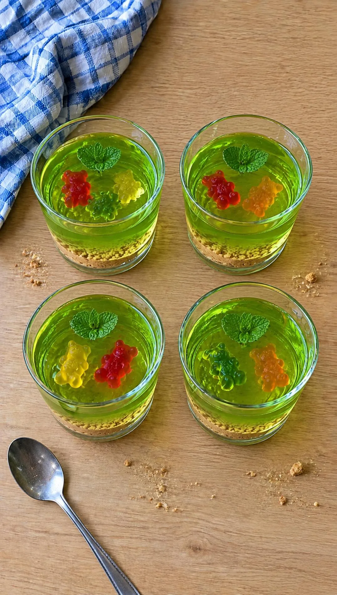 Glas mit grüner Götterspeise, Gummibärchen und Minzblatt, am Boden eine Schicht Keks-Sand