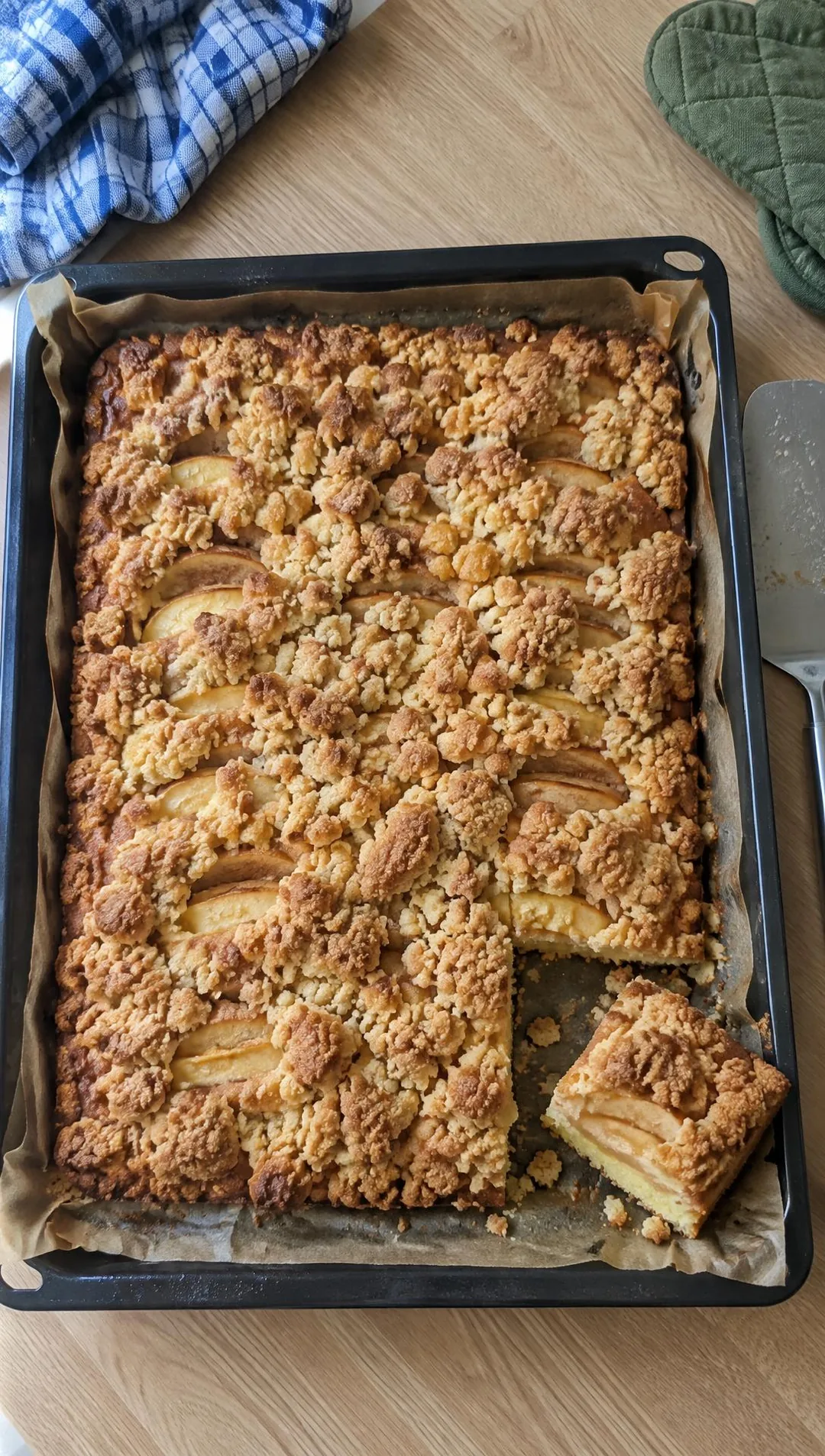Ein Pfannenwender hebt ein Stück Apfel-Streuselkuchen aus dem Blech, ein paar Streusel fallen herunter