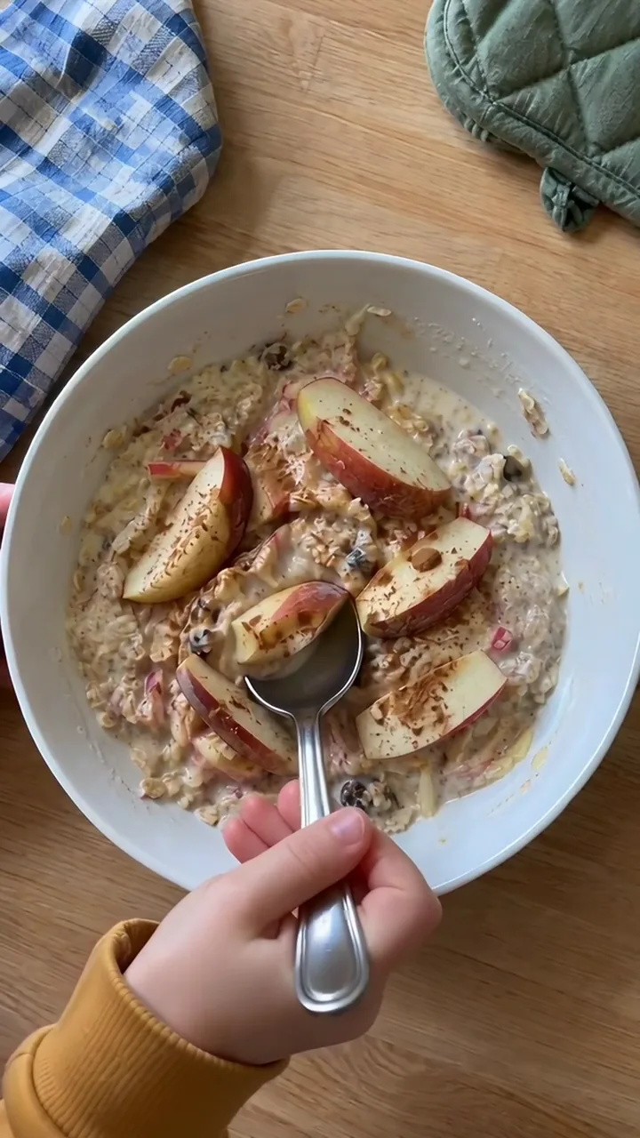 Fertige Müslischüssel, eine Kinderhand schöpft mit einem kleinen Löffel etwas cremiges Müsli mit Apfel heraus