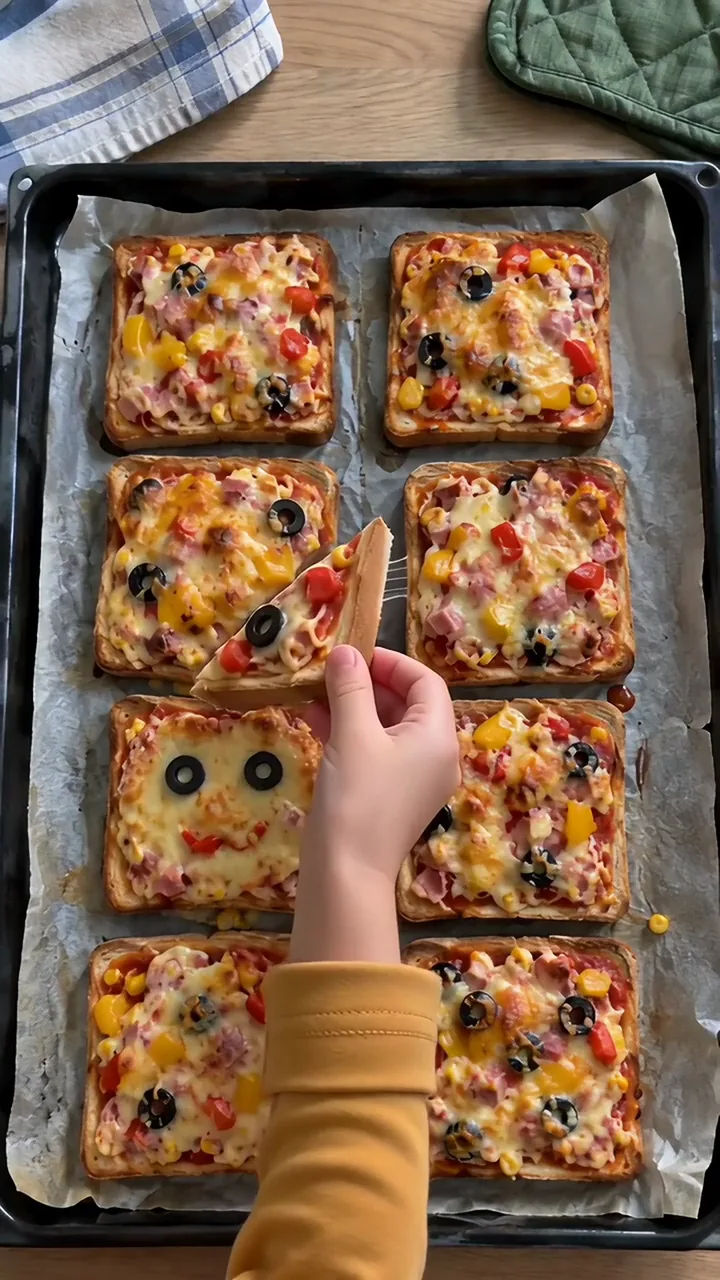 Eine Kinderhand hebt eine halbierte Toast-Pizza mit Olivenscheibe vom Blech, der Käse zieht Fäden