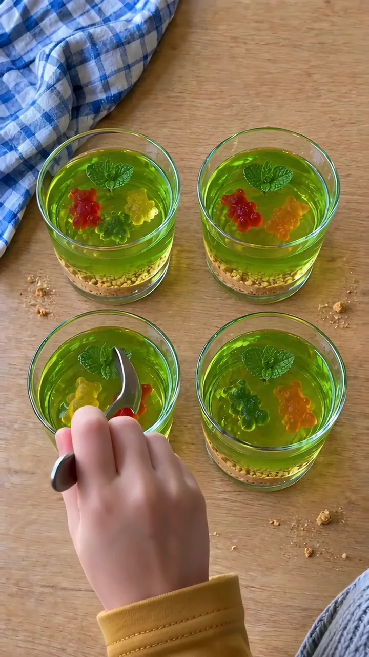 Eine Kinderhand löffelt aus einem Glas grüne Götterspeise mit Gummibärchen, Minzblatt und Keks-Sand am Boden