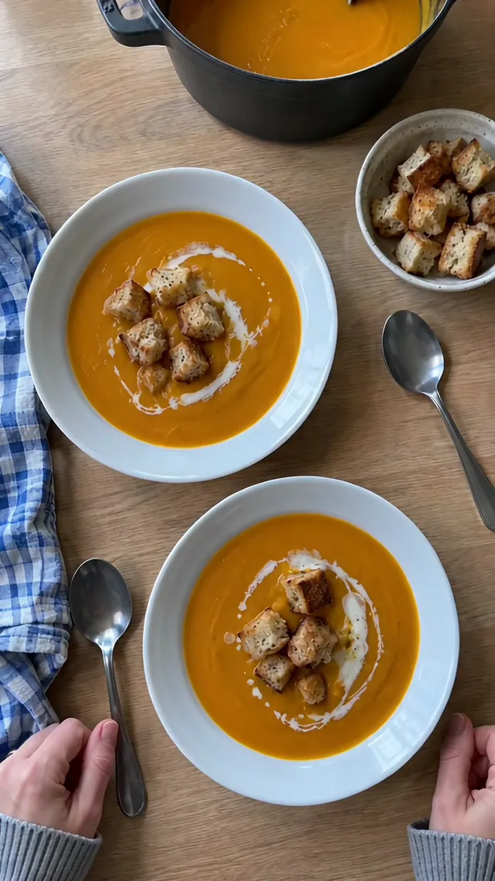 Kinderhand streut goldbraune Croûtons auf einen Teller mit orangefarbener Kürbissuppe