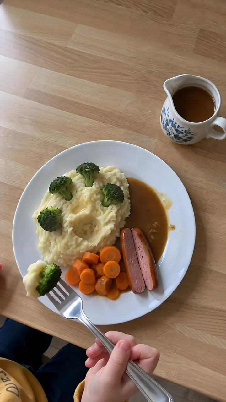 Eine Kinderhand nimmt mit der Gabel ein Brokkoli-Bäumchen mit Kartoffelbrei vom fertigen Teller