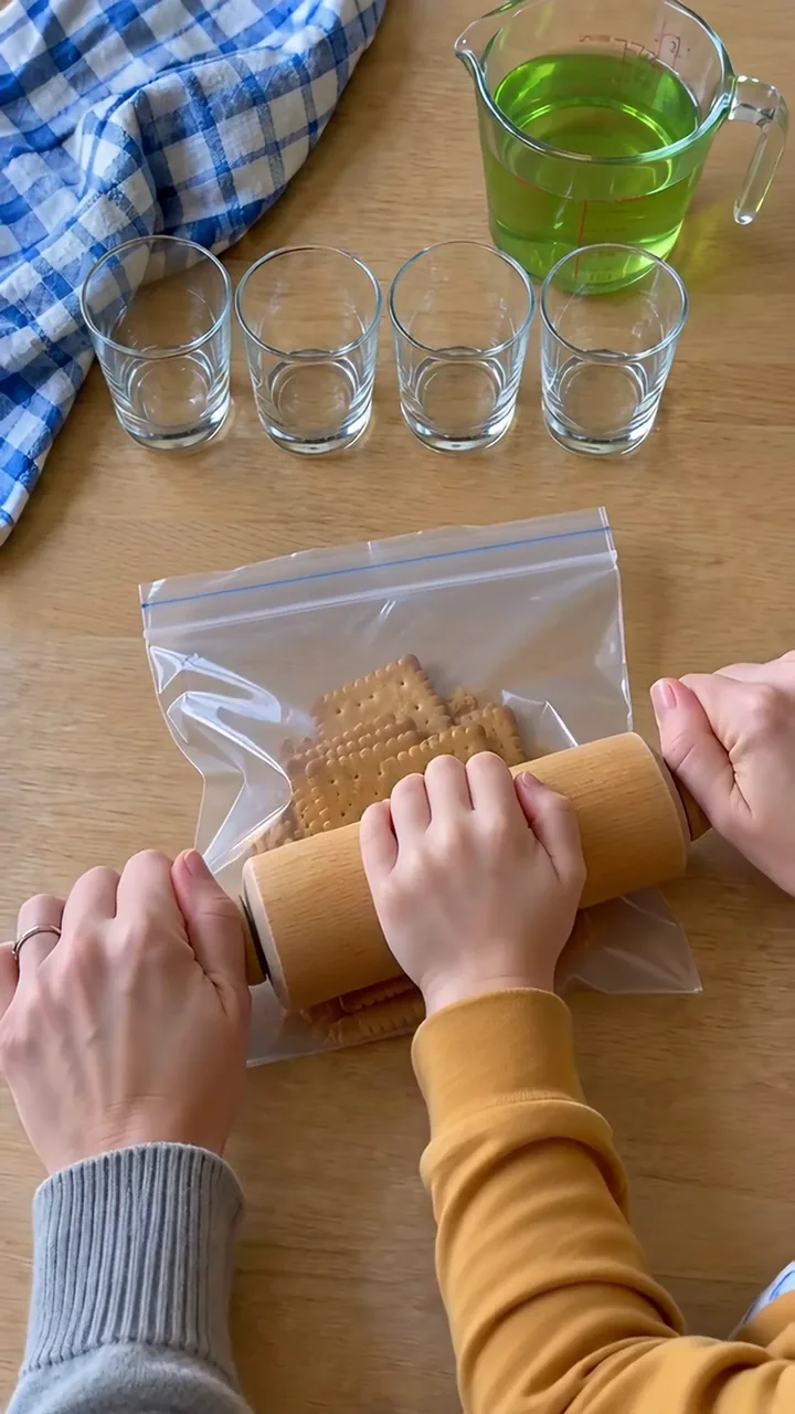 Ein Kind rollt mit dem Nudelholz über Butterkekse in einem Gefrierbeutel, ein Erwachsener hält den Beutel fest; dahinter vier leere Gläser und ein Messbecher mit grüner Götterspeise