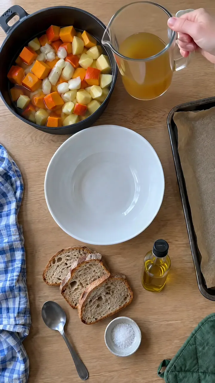 Erwachsenenhand gießt Gemüsebrühe über gewürfelten Kürbis, Kartoffeln und Zwiebel im Topf