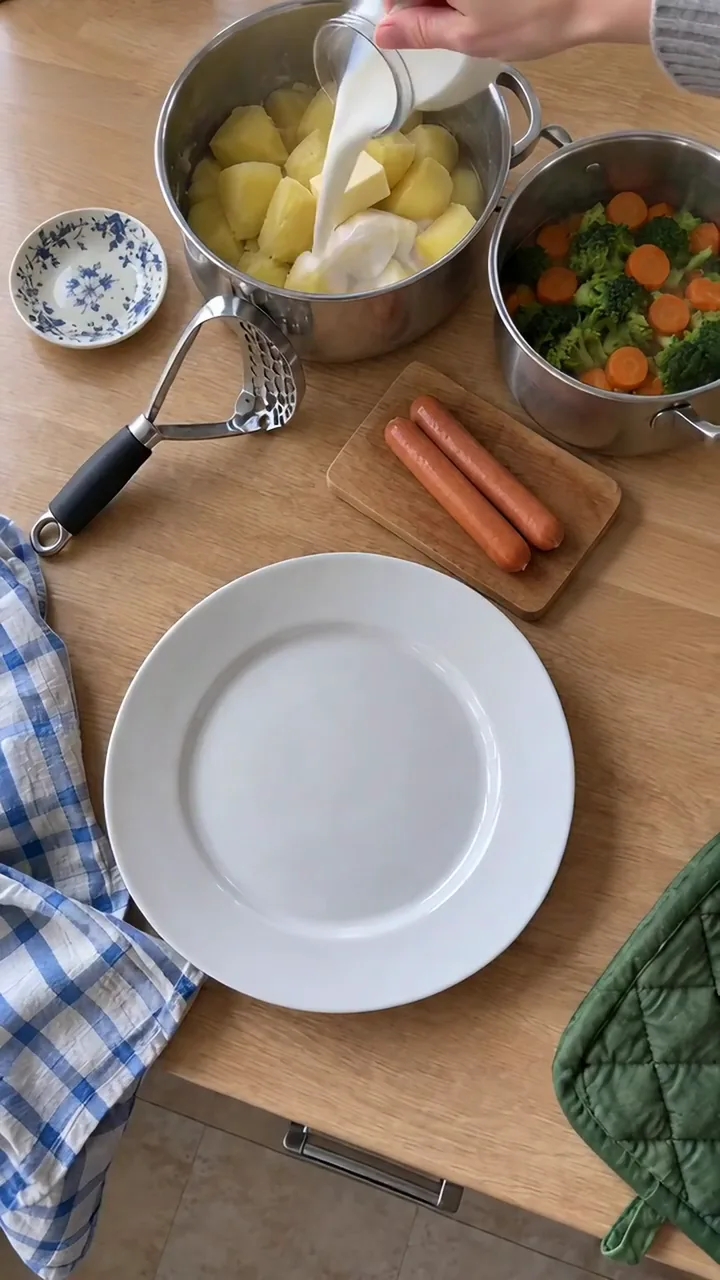 Warme Milch wird aus einem Glaskännchen über gekochte Kartoffeln und Butter im Topf gegossen, daneben steht ein Topf mit Brokkoli und Möhren
