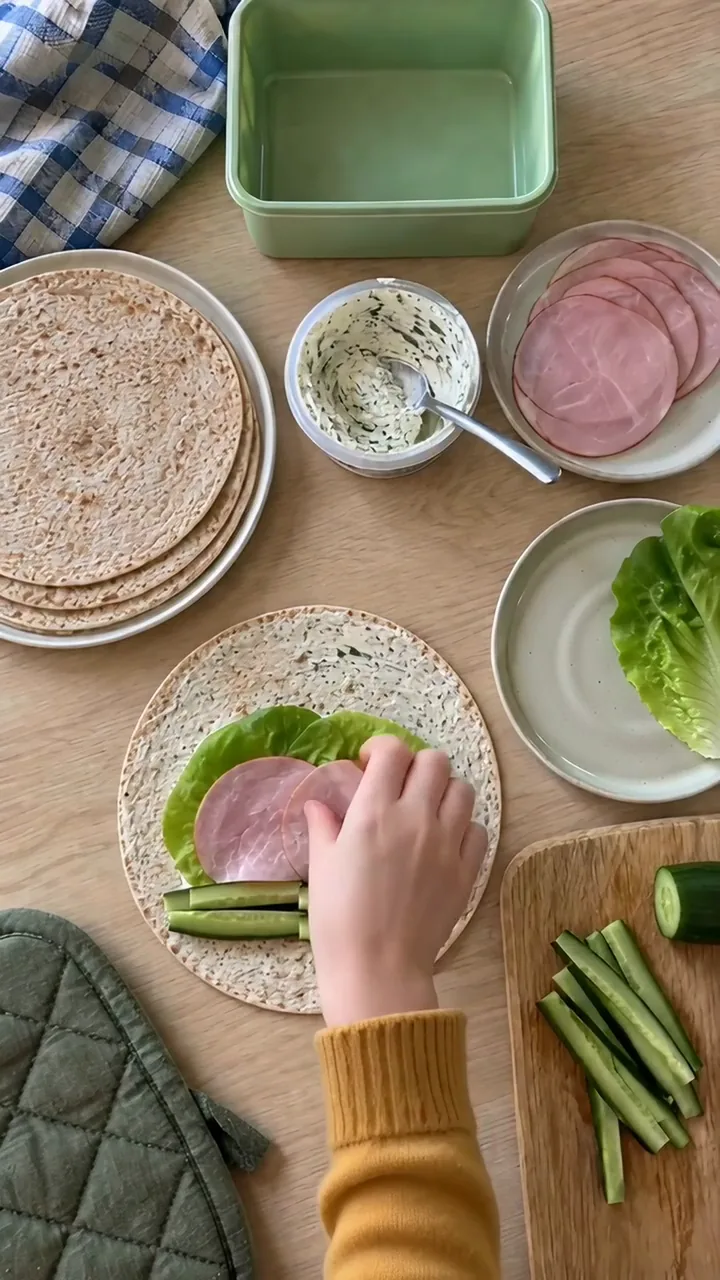 Kinderhände legen ein Salatblatt und Schinkenscheiben flach auf die bestrichene Tortilla