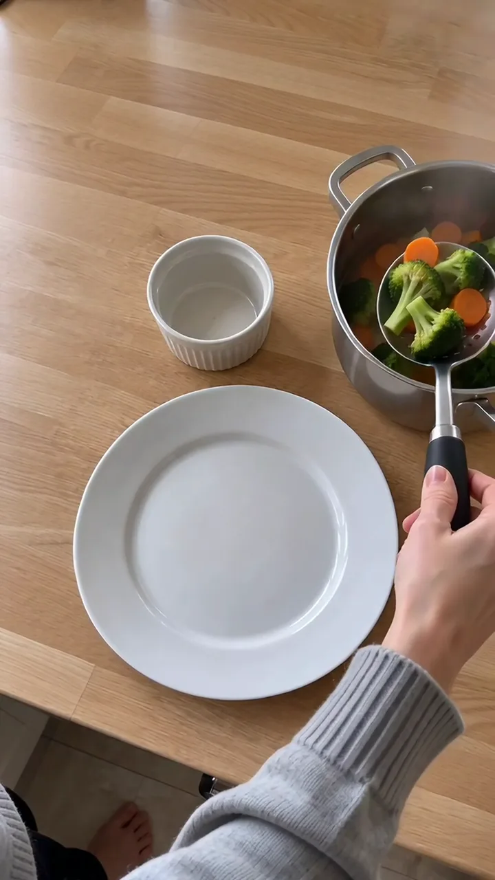 Eine Erwachsenenhand hebt mit einer Schaumkelle Brokkoliröschen und Möhrenscheiben aus dem Topf