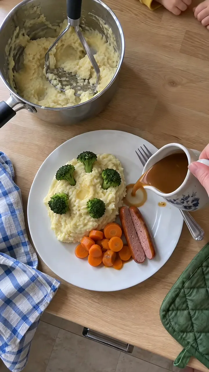 Aus einem Kännchen wird Bratensoße neben den Kartoffelbrei mit Brokkoli-Bäumchen, Möhren und einem halbierten Würstchen gegossen