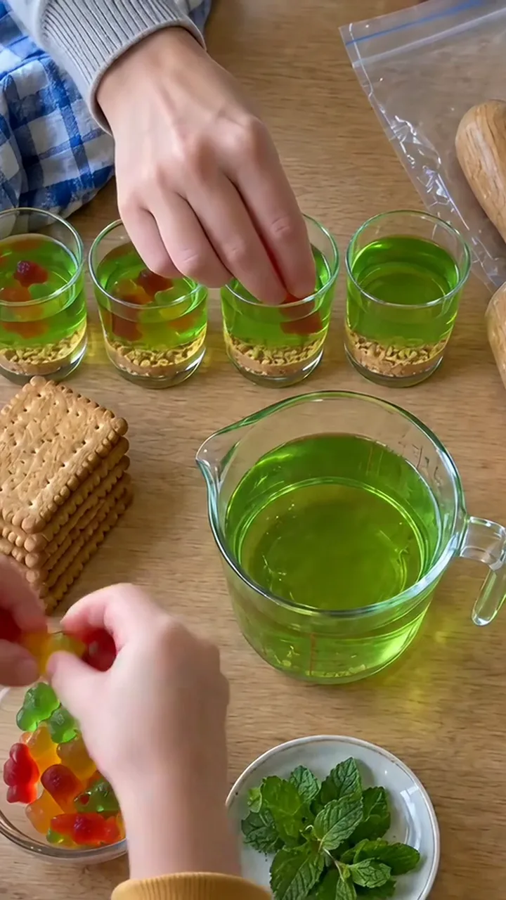 Gummibärchen werden in die halbfeste grüne Götterspeise in den Gläsern gedrückt; eine Kinderhand hält weitere Gummibärchen
