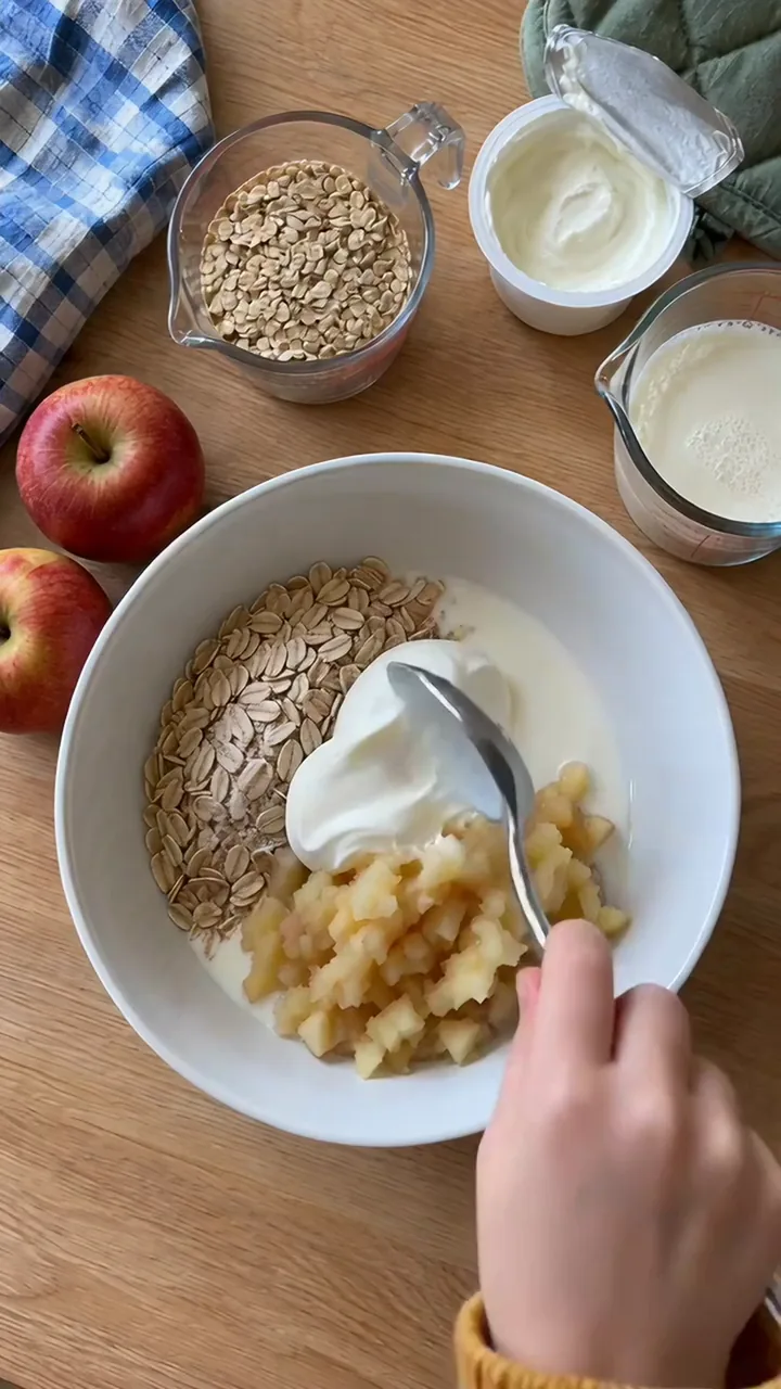 Kinderhand rührt Haferflocken, Joghurt, Milch und geriebenen Apfel mit einem großen Löffel zu einer cremigen Masse