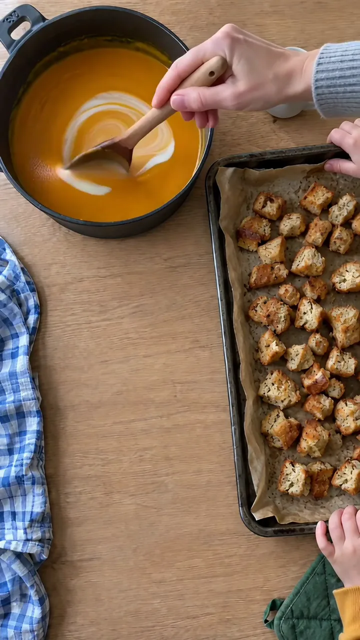 Erwachsenenhand gießt Sahne in die glatte orangefarbene Suppe und rührt sie mit einem Kochlöffel unter